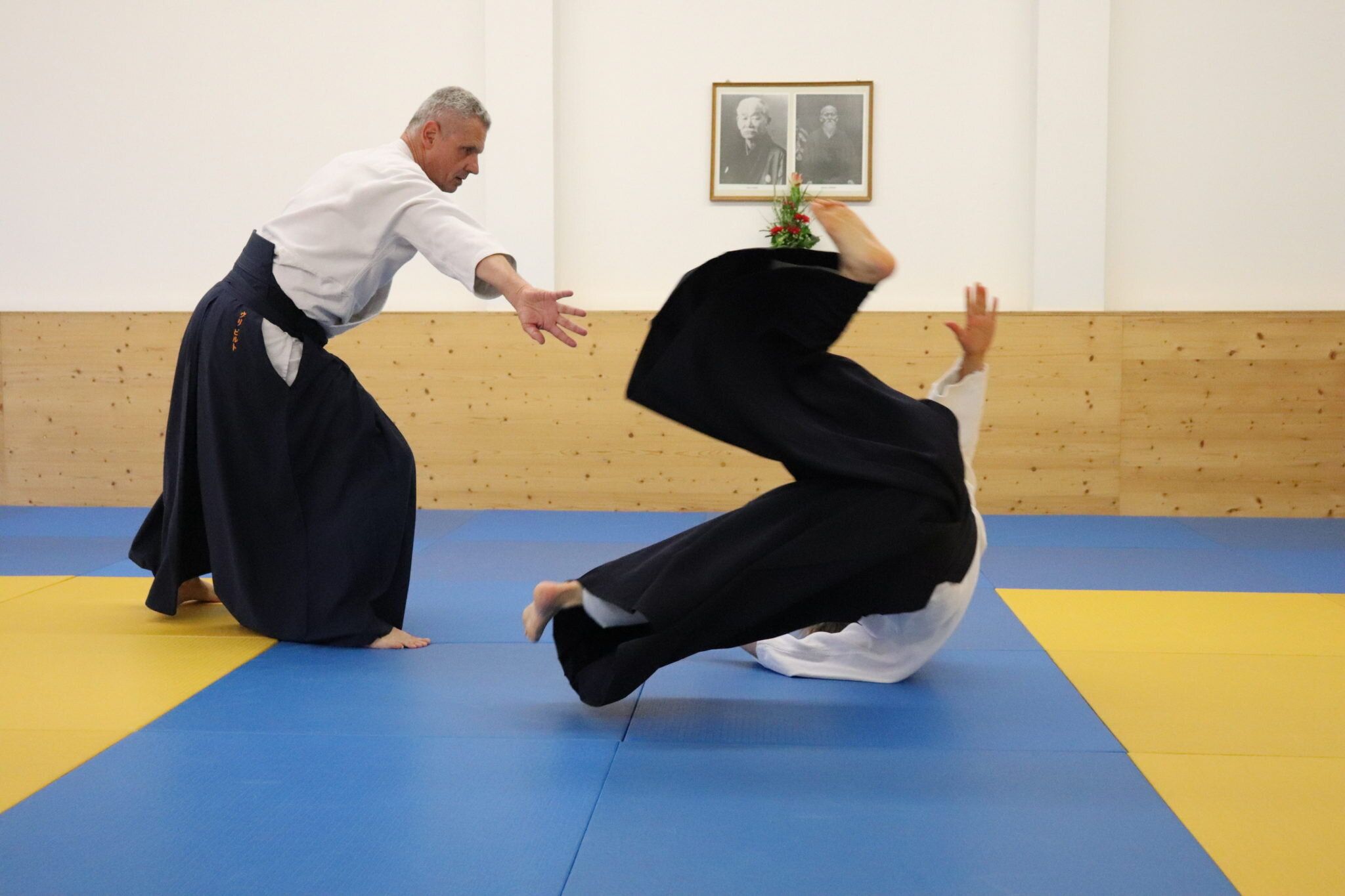 Zwei Aikidoka in Hakama im Dojo: Der Werfende führt mit ausgestrecktem Arm, der Angreifer dreht sich in einer Rolle durch die Luft; im Hintergrund die Bilder der Gründer.